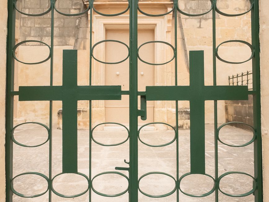 Green iron gate featuring cross design, in front of historical stone building entrance.